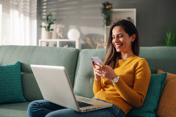 Portrait of a woman using laptop and a phone while sitting on a couch at home - obrazy, fototapety, plakaty