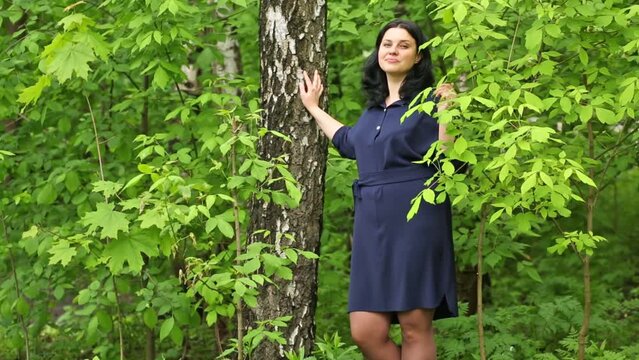 Woman in blue dress stand near birch and smell leaf in park.