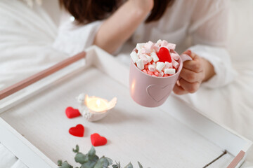 Beautiful woman with pink mug of cocoa, marshmallows and heart-shaped candy on bed at home. St Valentine's Day.