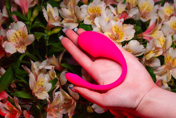 Pink vaginal vibrator for the G-spot in a girl’s hand against a background of flowers. Sex Shop...
