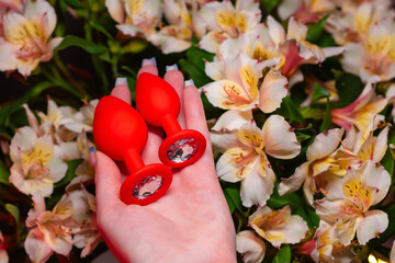 Two red silicone anal plugs in a girl’s hand against a background of flowers. Sex Shop Products,...