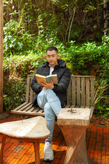 Fototapeta premium Asian young man sitting on a wooden bench reading a novel in a park