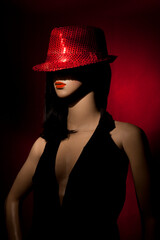 Plastic woman mannequin with clack long hair, red lipstick, and wearing a bright red sequins hat posing on a red background in a one light studio setting