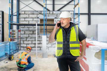 Man builder. Foreman in room under renovation. Builder guy is talking on phone. Repairman in reflective vest and helmet. Foreman standing in building under construction. Man architect calling client