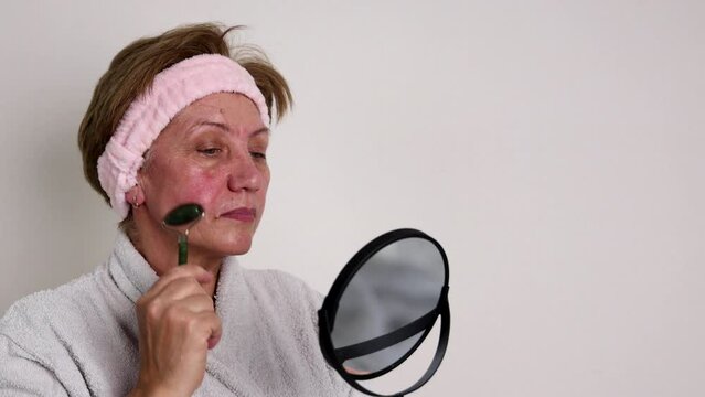 elder women using gua sha and jade roller for facial massage. anti aging, wrinkles, rejuvenation procedures. grandmother with gray hair with headband, in bathrobe, holding mirror in hand.old lady