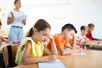 Young girl and boy studying subject in classroom, listening to teacher and writing.
