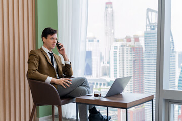 Closeup of handsome businessman making phone call with manager while sitting near window with skyscraper view. Executive manager talking working by using phone and laptop. Look aside. Ornamented.