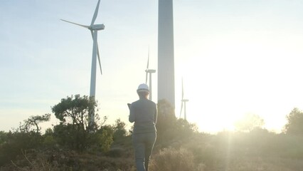 Female engineer with electronic tablet and portable device inspects wind energy installation and turbines during sunset super wide angle shot and moving camera