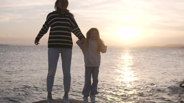 Beautiful mom and little daughter dancing on a rock by the sea at sunset,