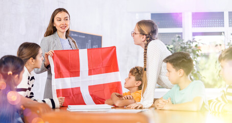 Joyful young woman teacher demonstrating flag of Denmark for schoolkids preteens during lesson of...
