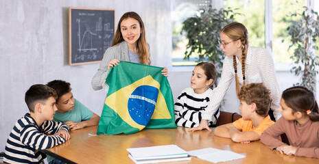 Positive young female teacher holding national flag of Brazil while working with preteens schoolgirls and schoolboys in classroom