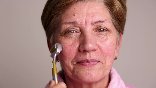 portrait head shoot of beautiful elder woman using derma roller with needles for facial rejuvenation, face anti aging wrinkle procedures at home. smiling female granny