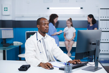 Fototapeta premium Dedicated African American male doctor using the desktop pc to document his medical notes. In the background, a caucasian nurse attends to a child in a well-equipped examination room.