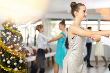Smiling elegant teenage girl dancing slow waltz as couple with classmate during festive Christmas...