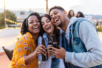 Close-up frontal image of three multi-ethnic young adult friends embracing and drinking beer in an outdoor terrace while celebrating a party. Celebrating together and diversity - obrazy, fototapety, plakaty