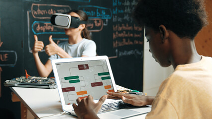 Caucasian school girl wear VR glass and showing thumb up while african boy working on laptop at...