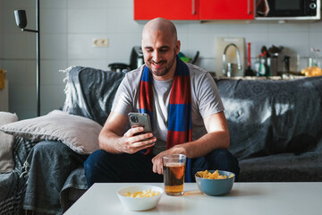 Young man looking at the phone smiling while watching sports on the sofa