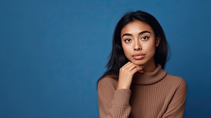 Thoughtful Asian woman, keeps her hand on her chin, looks thoughtful, dressed in a casual blue sweater, worried expression.