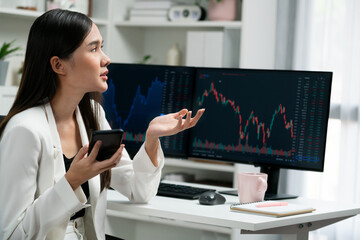 Obraz na płótnie Canvas Stressful young beautiful Asian businesswoman focusing on smartphone, analyzing possible risk to profit or loss in dynamic exchange stock market graph on pc screen at modern office. Stratagem.