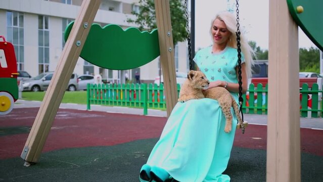 Blonde sits on swing and pats lion calf which lies on her legs