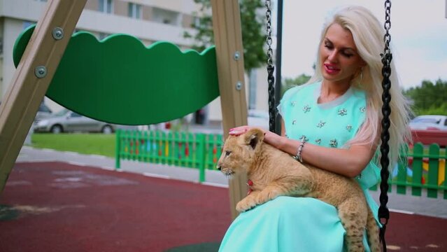 Woman sits on swing and pats lion cub which lies on her legs