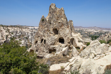 Sunny landscape of Cappadocia&rsquo;s unique fairy chimneys with cavities, surrounded by green shrubs, under a clear blue sky, highlighting nature&rsquo;s artistry.