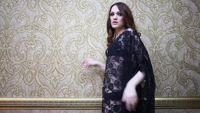 Brunette in black transparent dress poses near wall with expensive wallpapers