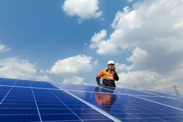 Male technician in work clothes while using radio communications standing near solar panels against a blue, cloudy sky.