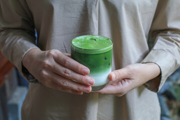 Female hand holding a glass of ice matcha green tea latte  in a beautiful minimal cafe. Organic...