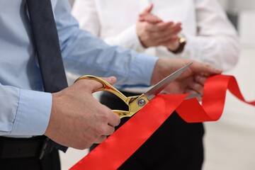 Man cutting red ribbon with scissors on blurred background, closeup