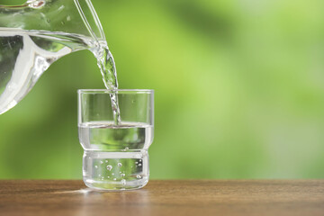 Pouring fresh water from jug into glass on wooden table against blurred green background, closeup....