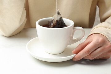 Woman putting tea bag in cup with hot water at white table, closeup