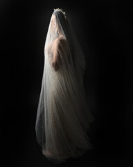 Full length portrait of beautiful model wearing white wedding gown and ghostly flowing veil like a shroud. Moody cinematic lighting, isolated on a dark studio background.