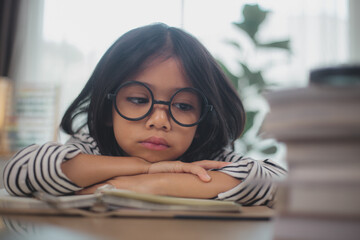Little Asian girl sitting alone and looking out with a bored face, Preschool child laying head down on the table with sad bored with homework, spoiled child - obrazy, fototapety, plakaty