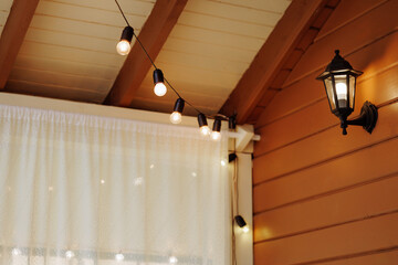 A cozy wooden patio is adorned with warmly lit string lights and a classic lantern. The front view...
