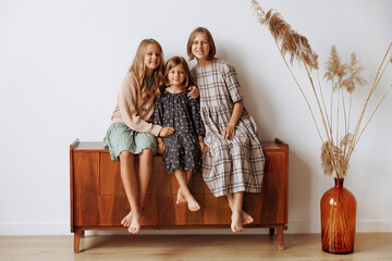Three Caucasian girls in home dresses of beige and brown colors are sitting on a wooden couch...