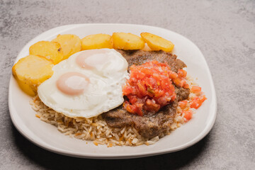 Authentic, traditional Bolivian meal: Silpancho, made of beef, potatoes, rice, fried eggs, tomato and onion salad. close-up on white plate on gray background. copy space