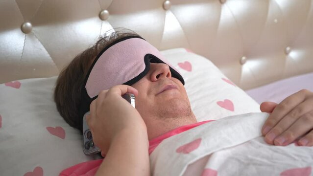Young man in sleep mask lies on double bed and talks on contemporary smartphone. Drowsy guy throws mobile phone on bed and continues sleeping