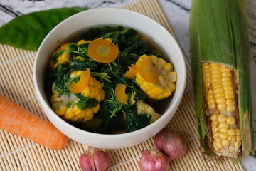 Sayur Bening Bayam is spinach clear soup, made of fresh spinach, carrot, corn, red onion, bay leaf. This soup served in white bowl. Indonesian food. Top view, flat lay, wooden table. 