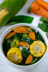 Sayur Bening Bayam is spinach clear soup, made of fresh spinach, carrot, corn, red onion, bay leaf. This soup served in white bowl. Indonesian food. Top view, flat lay, wooden table. 