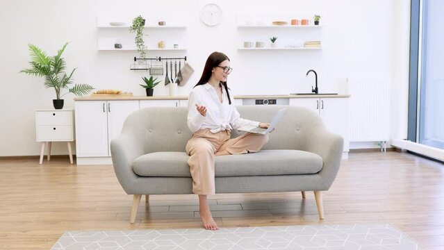 Positive brunette young female in casual attire gesturing with hands while sitting on couch with modern laptop and looking at web camera. Concept of unification people and technology.
