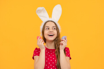 Teenager child wearing bunny ears and hold colorful painted easter eggs isolated at yellow...