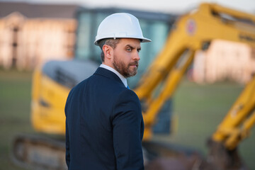 Construction owner near excavator. Confident construction owner in front of house. Architect, civil engineer. Man construction owner with a safety vest and hardhat at construction site.