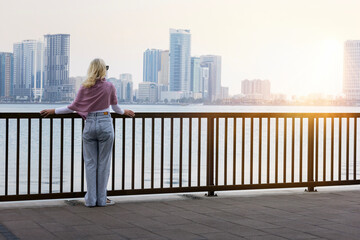 mature woman traveller looking out over promenade of Sharjah, UAE enjoying the gentle breeze and the beautiful cityscape at dusk.