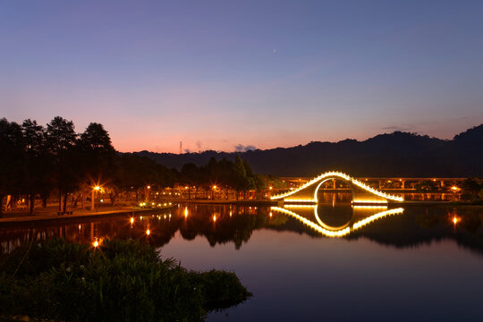 Early morning scene of a suburban park under twilight sky, with an arch bridge of Chinese style illuminated by lights & reflected in smooth lake water in Dahu Community Park Neihu, Taipei City, Taiwan