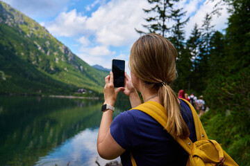Back view of woman hiking near mountain lake with backpack, taking photo with smartphone. Female...