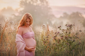 Fototapeta na wymiar A pregnant girl with long red hair looks at her belly. At dawn. Summer fog. Tenderness. Pregnancy. Future mom. Early morning. Sunrise. Summer. Belly of a pregnant woman. Beautiful mother. Family. 9 mo