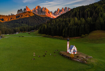 Val Di Funes, Dolomites, Italy - Aerial view of the beautiful St. Johann Church (Chiesetta di San Giovanni in Ranui) at South Tyrol with cows and Italian Dolomites in warm sunset colors at background