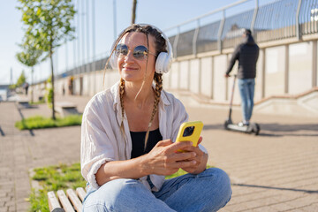 hipster woman in bluetooth headphones enjoying positive music playlist from mobile radio using 4g wireless in city, have video call, girl holding coffee to go and smartphone.