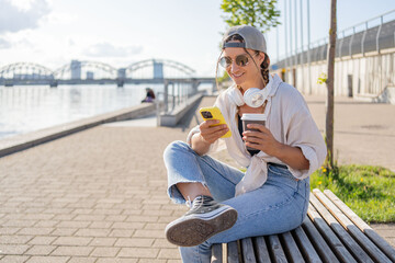 Fototapeta na wymiar Young Hispanic hipster girl browsing mobile phone while sitting in street with takeaway beverage. 